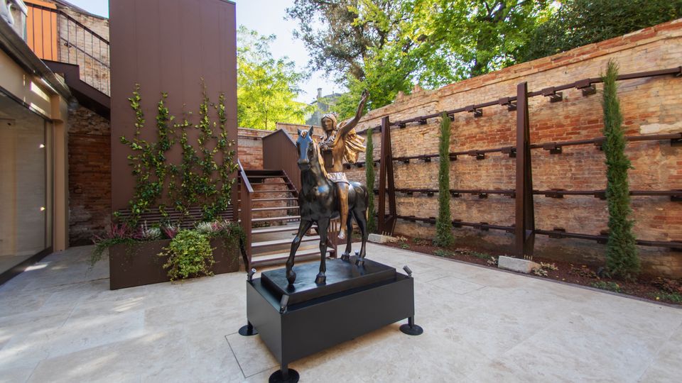 Cortile interno pavimentato con statue in bronzo su piedistallo al centro, circondato da muri in mattoni a vista e strutture metalliche moderne. Sullo sfondo si vedono scale, fioriere con rampicanti e alberi che ombreggiano lo spazio.