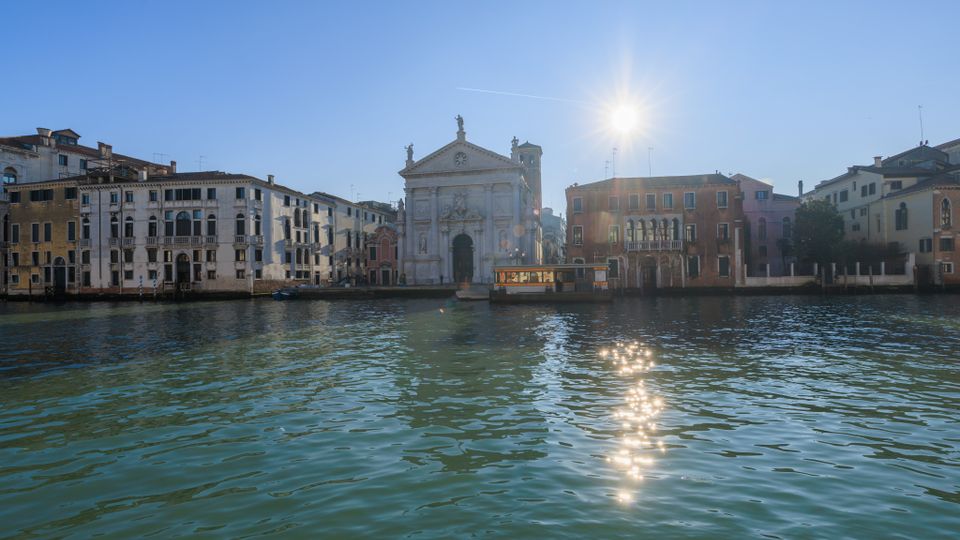 Veduta di un canale urbano con una chiesa in pietra chiara al centro e palazzi storici a più piani lungo le rive. In primo piano l’acqua del canale con riflessi del sole e un pontile per vaporetti davanti alla chiesa.