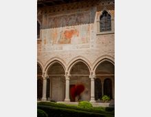 Chiostro in mattoni con portico ad archi gotici sostenuti da colonne in pietra e un giardino con siepi potate in primo piano. Sulla parete superiore si vede un grande affresco medievale sbiadito e una finestra ogivale con grata decorativa.