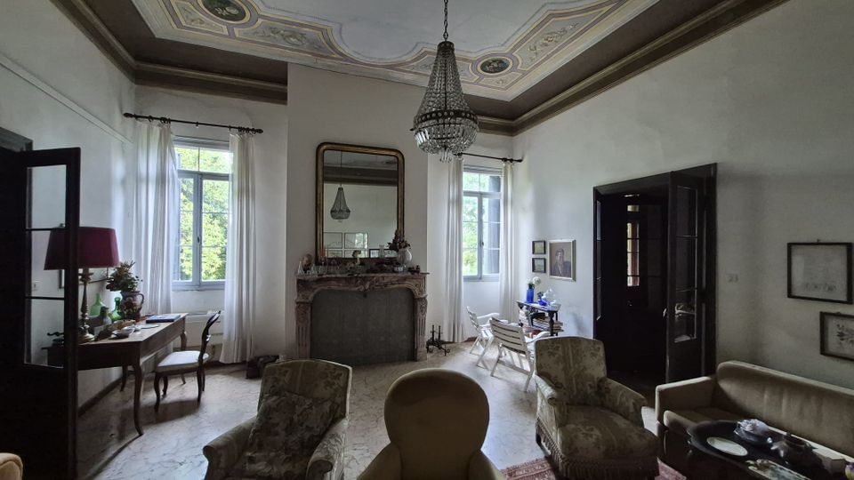 Salone di una casa d’epoca con soffitto decorato a stucchi e lampadario a gocce, camino in marmo con grande specchio e ampie finestre con tende bianche. Arredi classici con poltrone e divani, tavolini, scrivania e porte scure su pavimento chiaro.
