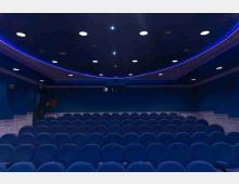 Interno di una sala cinema con file di poltrone blu disposte in platea. Soffitto scuro con faretti circolari incassati e una striscia luminosa blu che segue una curva lungo il bordo.