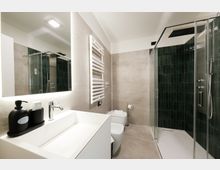 Bagno moderno con lavabo bianco e grande specchio illuminato, pareti rivestite in piastrelle grigie e scaldasalviette verticale. A destra una doccia con box in vetro e rivestimento in piastrelle verde scuro, con soffione a pioggia.