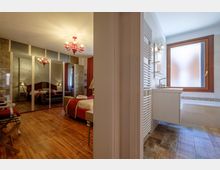 Interno di una suite con camera da letto elegante a pavimento in legno, armadi a specchio e lampadario rosso, affacciata su un bagno moderno con lavabo sospeso, termoarredo e vasca sotto una grande finestra satinata.