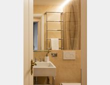 Bagno moderno rivestito in pietra beige, con lavabo squadrato bianco, mensola in vetro e scaldasalviette metallico a parete. Sulla destra si vede la parete della doccia con venature verticali e il pulsante di scarico incassato.