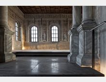 Interno di un edificio storico con grandi colonne in pietra e pavimento in lastre scure, con una pedana rialzata al centro. Sul fondo si vedono alte finestre ad arco che illuminano pareti affrescate con nicchie e statue, sotto un soffitto ligneo a cassettoni.