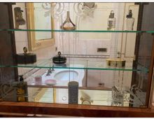 Mobiletto da bagno con ripiani in vetro e decorazioni incise, contenente flaconi e accessori da toilette. Sullo sfondo si vede un lavabo incassato con rubinetteria in metallo e pareti piastrellate chiare con dettagli dorati.