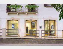 Facciata di un edificio con grandi vetrine al piano terra che mostrano un interno illuminato tipo ufficio o negozio, con cornici in pietra attorno alle aperture. Sopra, un balcone con ringhiera in ferro battuto e fioriere verdi, e una rampa esterna con corrimano davanti all’ingresso.