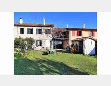 Cortile erboso di una casa di campagna con due edifici in muratura e tetti in coppi, persiane colorate e un piccolo annesso con portico. Nel prato si vedono un albero e una struttura tipo rete da gioco, con cielo sereno sullo sfondo.