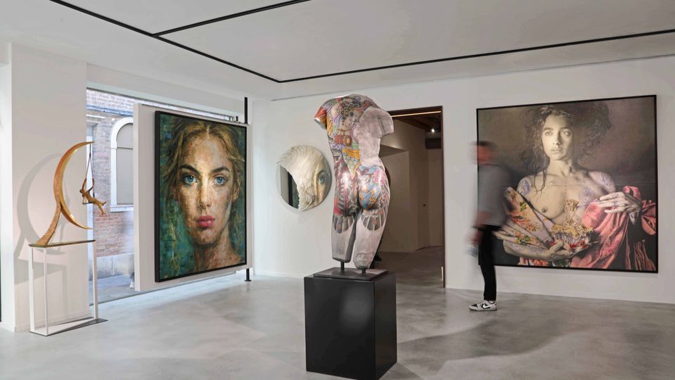 Interno di una galleria d’arte contemporanea con pareti bianche e pavimento grigio, illuminata da un soffitto a pannelli. Nella sala sono esposti grandi quadri figurativi alle pareti e una scultura su piedistallo al centro, con una vetrata laterale che affaccia su un edificio in mattoni.