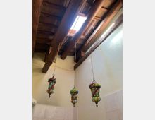 Interno con soffitto alto in legno a travi a vista e lucernario rettangolare che lascia entrare luce naturale. Tre lanterne sospese in vetro colorato e struttura in ferro pendono da lunghi cavi sopra pareti intonacate chiare.