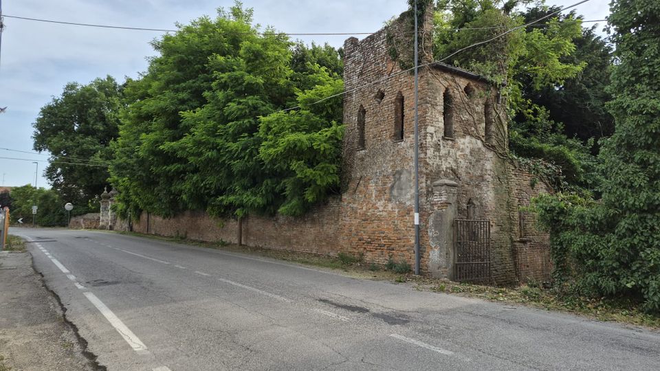 Strada asfaltata di campagna che costeggia un alto muro in mattoni e un edificio in rovina con piccole finestre ad arco. La struttura è parzialmente coperta da edera e alberi fitti, con un cancello metallico e un lampione vicino all’angolo.