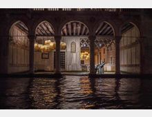 Cortile coperto con portico ad archi e colonne in pietra affacciato su un canale, con acqua in primo piano che riflette le luci. Sullo sfondo si vedono pareti in legno e pietra, una grata decorativa e un lampadario artistico illuminato.