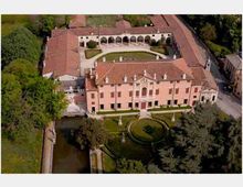 Veduta aerea di una villa storica con facciata rosa e tetto in coppi, affacciata su un giardino formale con aiuole geometriche e statue. Sul retro si vede un grande cortile con porticato ad archi, circondato da edifici annessi e alberi, con un canale lungo il lato sinistro.