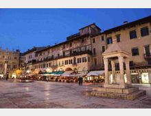 Piazza lastricata al crepuscolo con un piccolo tempietto in pietra su basamento in primo piano. Sullo sfondo si vede una lunga fila di edifici storici con portici, finestre e balconi, e caffè con ombrelloni lungo il lato della piazza.