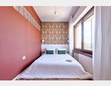 Camera da letto stretta con letto matrimoniale rivestito di coperta bianca e due cuscini verdi, parete laterale color terracotta e parete di fondo con carta da parati decorata e boiserie bianca. A destra, grande finestra con telaio in legno e tende bianche che lascia entrare molta luce naturale.