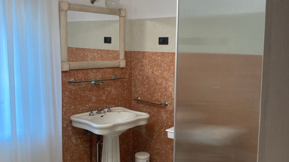 Bagno interno con lavabo su piedistallo e specchio con cornice decorata, pareti rivestite a metà in piastrelle marmorizzate color terracotta e parte superiore bianca. Pavimento a motivi geometrici con stelle e, a destra, una parete divisoria in vetro satinato rigato vicino alla zona vasca o doccia.