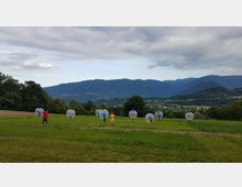 Ampio prato in collina con sullo sfondo una valle e una catena montuosa sotto un cielo nuvoloso. Nel campo sono visibili grandi sfere trasparenti distanziate lungo una linea, con alberi ai margini e terreni agricoli nelle vicinanze.
