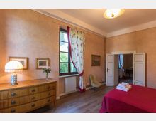 Camera da letto in stile classico con pareti color pesca, pavimento in legno e letto con copriletto rosso. Grande finestra con tende a motivo floreale, cassettiera con lampada e quadri, poltroncina e porta bianca che conduce a un’altra stanza.