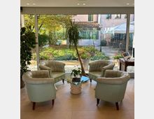 Salotto luminoso con quattro poltrone imbottite color crema disposte attorno a un tavolino in vetro, davanti a una grande vetrata a tutta parete. All’esterno si vede un giardino fiorito con vialetto e una terrazza con tavoli sotto una tenda, accanto a un edificio con tetto in coppi.