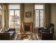 Salotto elegante con due grandi finestre a tutta altezza con tende chiare, che affacciano su edifici storici. Al centro ci sono un camino in marmo rosato con orologio a parete sopra, un tavolino in vetro su tappeto e divani e poltrone in pelle scura.