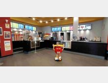 Interno di un cinema con area snack bar e biglietteria: lungo bancone nero con vetrine, macchine per popcorn e bevande, e schermi illuminati con il menu sulla parete. Al centro della sala, su pavimento in moquette, c’è una statua promozionale rossa a forma di caramella con vassoio.