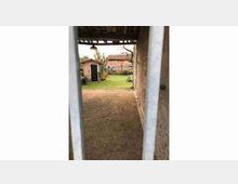 Passaggio coperto in una struttura rustica con pareti in muratura e pavimento in ghiaia, che conduce a un cortile erboso. Sullo sfondo si vedono edifici in mattoni con tetti in tegole e alcuni alberi.