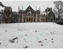 Facciata di un grande edificio storico in stile neogotico, con archi a sesto acuto, finestre alte e piccole torri laterali. Davanti si estende un ampio prato coperto di neve, con alberi spogli ai lati.