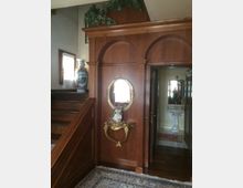 Interno di un'abitazione con pareti rivestite in legno scuro, un arco decorativo, uno specchio con cornice dorata e consolle sottostante. A sinistra si trova una scala in legno, e sullo sfondo è visibile un bagno con lavabo bianco.