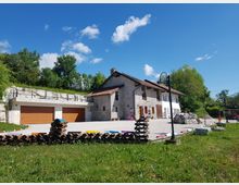 Cascina rurale ristrutturata con tetto in coppi e facciata chiara, affacciata su un ampio cortile in ghiaia con lampioni e piccoli elementi colorati lungo il bordo. A sinistra si vedono autorimesse con portoni in legno sotto un terrazzamento in pietra, circondati da prato e alberi.
