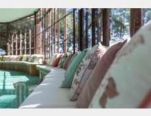 Veranda tipo serra con lunghe vetrate a telaio metallico affacciate su alberi, illuminata dalla luce naturale. Lungo la parete corre una panca curva con cuscini decorativi color pastello su pavimento lucido verde acqua.