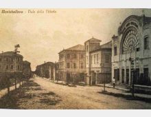Cartolina d’epoca di un ampio viale urbano a Montebelluna, con strada in terra battuta e filare di giovani alberi lungo il bordo. Ai lati si vedono edifici in muratura a più piani, tra cui sulla destra una grande facciata con arco e ampia vetrata semicircolare.