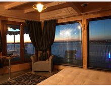 Interno di una camera in legno con soffitto a travi, lampada a sospensione, poltrona e letto, affacciata su grandi finestre e una porta finestra con tende e veneziane. Dalle vetrate si vede un balcone con ringhiera e una vista sul mare al tramonto.