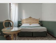 Camera da letto in stile classico con letto matrimoniale e testiera decorata, pareti bicolore verde e bianco e pavimento a mosaico. Una finestra con tende bianche illumina la stanza, con piccolo tavolo rotondo e sedia in primo piano e luci a parete ai lati del letto.