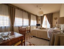 Camera da letto in stile classico con ampie finestre ad angolo coperte da tende chiare e drappeggi, letto matrimoniale con copriletto beige e comodini con lampade. Pavimento a scacchi, arredi in legno con scrivania e sedia, e un lampadario a sospensione al centro della stanza.