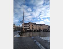 Ampia piazza urbana lastricata e bagnata dalla pioggia, con un alto pennone e un lampione in primo piano. Sullo sfondo si vedono edifici storici con portici ad archi e finestre scure, sotto un cielo azzurro con nuvole sparse.