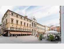 Ampia piazza cittadina con un grande edificio storico in stile rinascimentale, caratterizzato da facciata chiara con finestre ad arco e loggiato con colonne. Sul lato destro si vedono tavolini all’aperto sotto ombrelloni e piante in vaso, con altri palazzi colorati sullo sfondo.