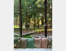 Vista da una grande finestra su un giardino alberato allestito con file di sedie in legno e cuscini bianchi, come per una cerimonia all’aperto. In primo piano, un divano con cuscini decorati e alcune sfere di vetro appese davanti al vetro.