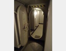 Porta stagna metallica con chiusura a ruota, aperta su un corridoio interno stretto con pareti grigie, illuminazione a parete e griglia di aerazione sul pavimento. Sopra il varco ovale sono visibili strisce di segnalazione giallo-nere e tubazioni lungo il soffitto.