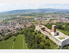 Veduta aerea di una fortezza in pietra su una collina, con mura perimetrali, cortile interno e una torre quadrata. Intorno si vedono vigneti e boschi, con un centro abitato e campi nella pianura sottostante e montagne sullo sfondo.
