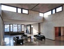 Ampio soggiorno in stile moderno con soffitto alto e pareti in intonaco grezzo, illuminato da finestre a nastro superiori e da grandi vetrate sul lato. Pavimento lucido e arredi minimal con divani in pelle nera e tavolino centrale in metallo.