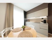 Cucina moderna e compatta con mobili bianchi a linea pulita, piano di lavoro con lavello e piano cottura illuminati da luce sotto pensile e parete marrone scuro. In primo piano un tavolo rotondo in legno con tovagliette intrecciate e sedie bianche, vicino a una grande finestra con tende chiare.