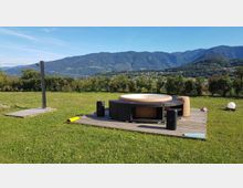 Vasca idromassaggio rotonda su una pedana in legno in un prato aperto, con un piccolo punto doccia esterno nelle vicinanze. Sullo sfondo si vedono colline e montagne boscose sotto un cielo sereno.