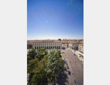 Veduta dall’alto di una grande piazza urbana con ampio incrocio stradale, fiancheggiata da edifici storici con facciate in pietra, arcate e finestre alte. In primo piano si vede un’area alberata con grandi chiome verdi e, sullo sfondo, i tetti della città sotto un cielo sereno.