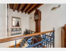 Pianerottolo interno con scala, soffitto a travi di legno a vista e pareti chiare. Ringhiera in ferro battuto decorato, grande orologio a pendolo a parete e passaggio verso un’altra stanza con specchio incorniciato.