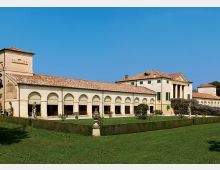 Complesso storico in stile villa con lunghi porticati ad archi e tetti in coppi, affacciato su un prato curato con siepi geometriche. Sullo sfondo si vede l’edificio principale con facciata classicheggiante e persiane verdi sotto un cielo sereno.