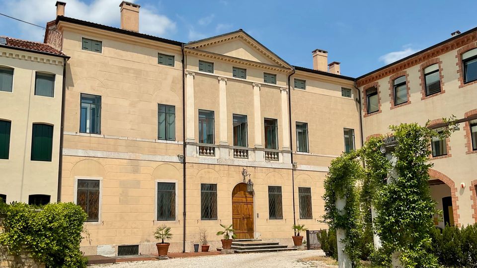 Facciata di una villa storica a due piani con intonaco chiaro, frontone centrale e quattro lesene che incorniciano un balconcino sopra un portale in legno. Davanti si vede un vialetto di ghiaia con vasi di piante e un pergolato con rampicanti nel cortile, tra edifici adiacenti.
