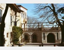 Cortile esterno di una villa storica intonacata, con portico ad archi al piano terra, terrazza con balaustra in legno e un piccolo balcone con arcate al piano superiore. Alberi spogli e vegetazione ai lati incorniciano la scena sotto un cielo sereno.
