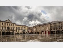 Ampia piazza lastricata bagnata dalla pioggia, con riflessi sul pavimento e cielo coperto da nuvole scure. Sullo sfondo si affacciano edifici storici con portici ad archi e facciate in pietra, tra cui un palazzo con orologio sulla sommità e un piccolo padiglione decorato.