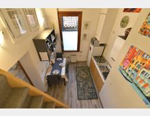 Cucina-soggiorno stretta e luminosa con parete attrezzata e tavolo da pranzo per quattro, vista dall’alto vicino a una scala interna. Sul lato destro c’è un angolo cottura lineare con fornelli e lavello, e in fondo una finestra alta con cornice in legno e termosifone sotto.