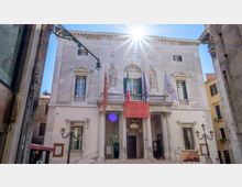 Facciata di un edificio storico in pietra chiara su una piccola piazza, con ingresso porticato a colonne, balcone centrale e nicchie con statue. Diverse bandiere e uno striscione pendono sopra l’entrata, con lampioni in ferro battuto e sole alto che crea un forte bagliore.